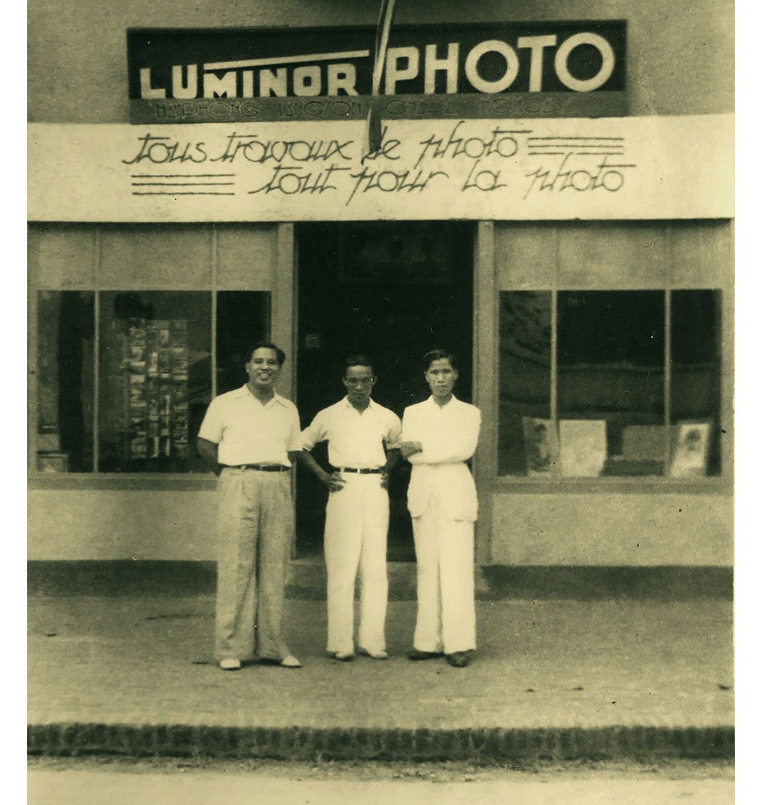 Ông Nguyễn Văn Chành (bìa phải) trước cửa tiệm ảnh với slogan bằng tiếng Pháp Ảnh: tư liệu triển lãm Những ngày đợi nắng